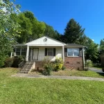 Exterior view of a small yellow ranch-style home with a green lawn and mature trees.